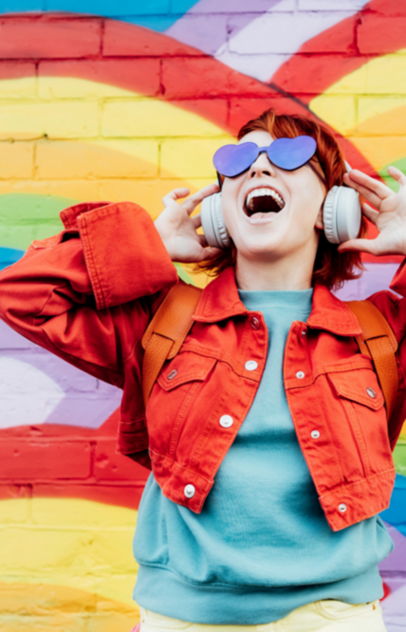 Frau steht lachend mit Kopfhörern vor einer bunten Wand